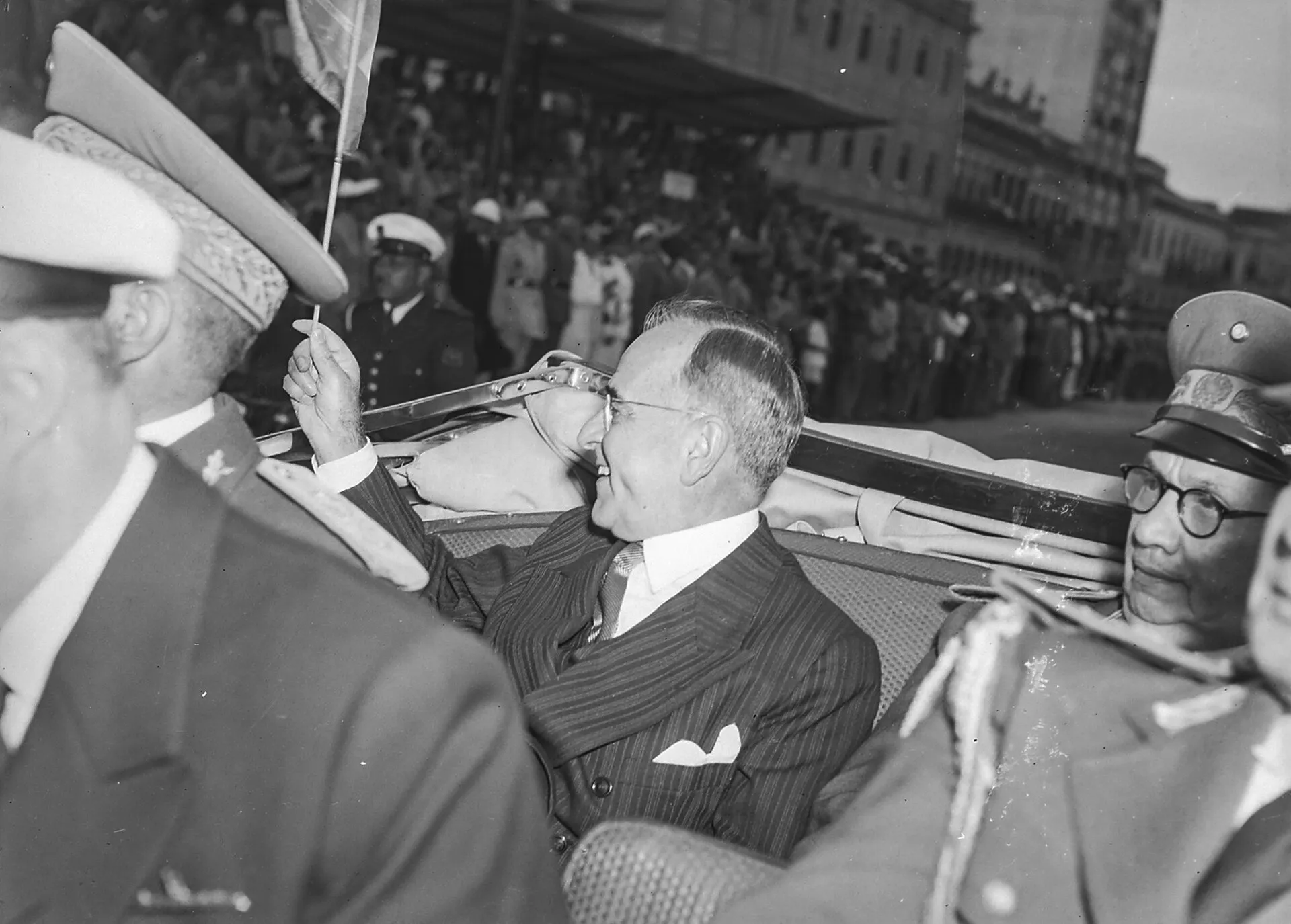Homem de terno listrado e óculos acena de um carro aberto, cercado por militares de quepe, durante um desfile no Rio de Janeiro em 1945