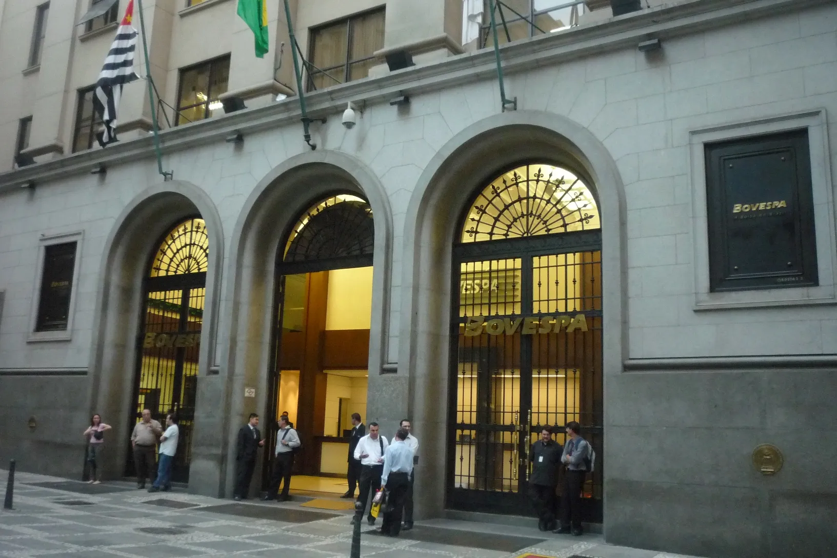Fachada de pedra da Bovespa, com três arcos de entrada e o letreiro BOVESPA, e pessoas na calçada, em São Paulo