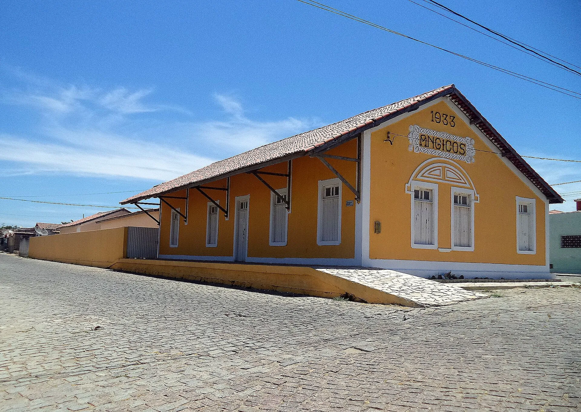 Antiga estação ferroviária de Angicos, prédio amarelo de telhado colonial com a inscrição Angicos e o ano 1933 na fachada, em rua de paralelepípedos