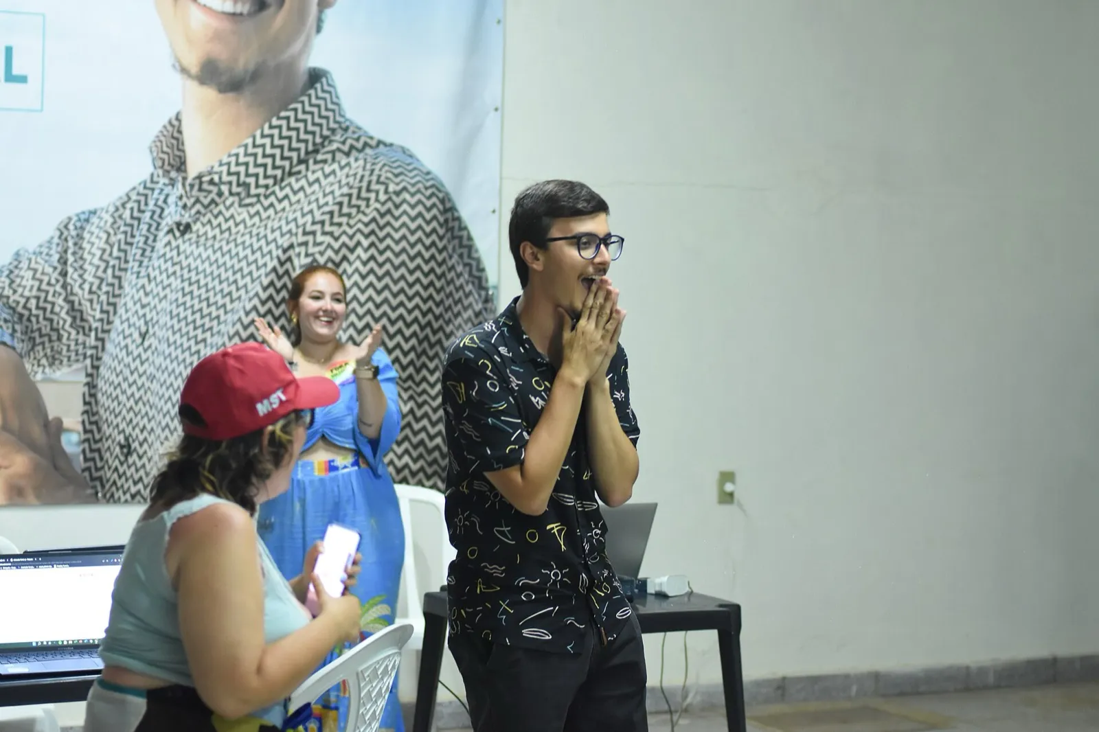 Homem de óculos reage com as mãos no rosto enquanto outras pessoas aplaudem em um local de acompanhamento da apuração da eleição de 2022, com um cartaz de campanha ao fundo