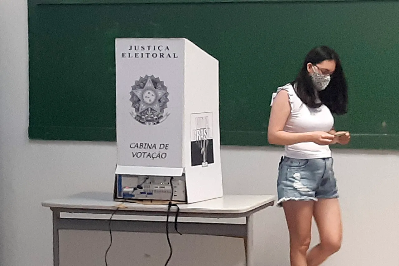 Eleitora de máscara ao lado de uma cabine de votação da Justiça Eleitoral, em sala de aula com quadro-negro verde ao fundo