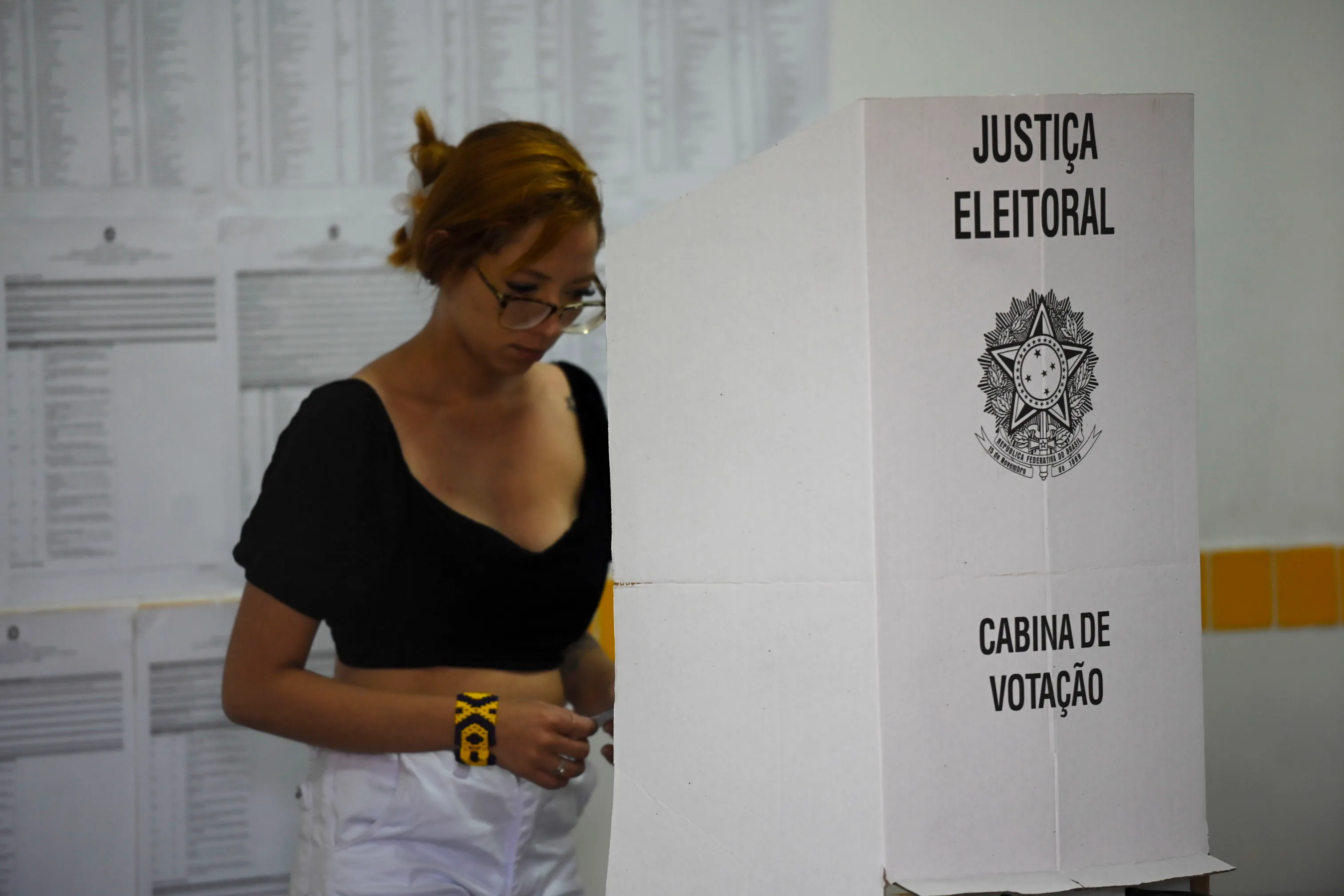 Eleitora de blusa preta e óculos entra em uma cabina de votação branca, com o letreiro Justiça Eleitoral, em uma seção eleitoral