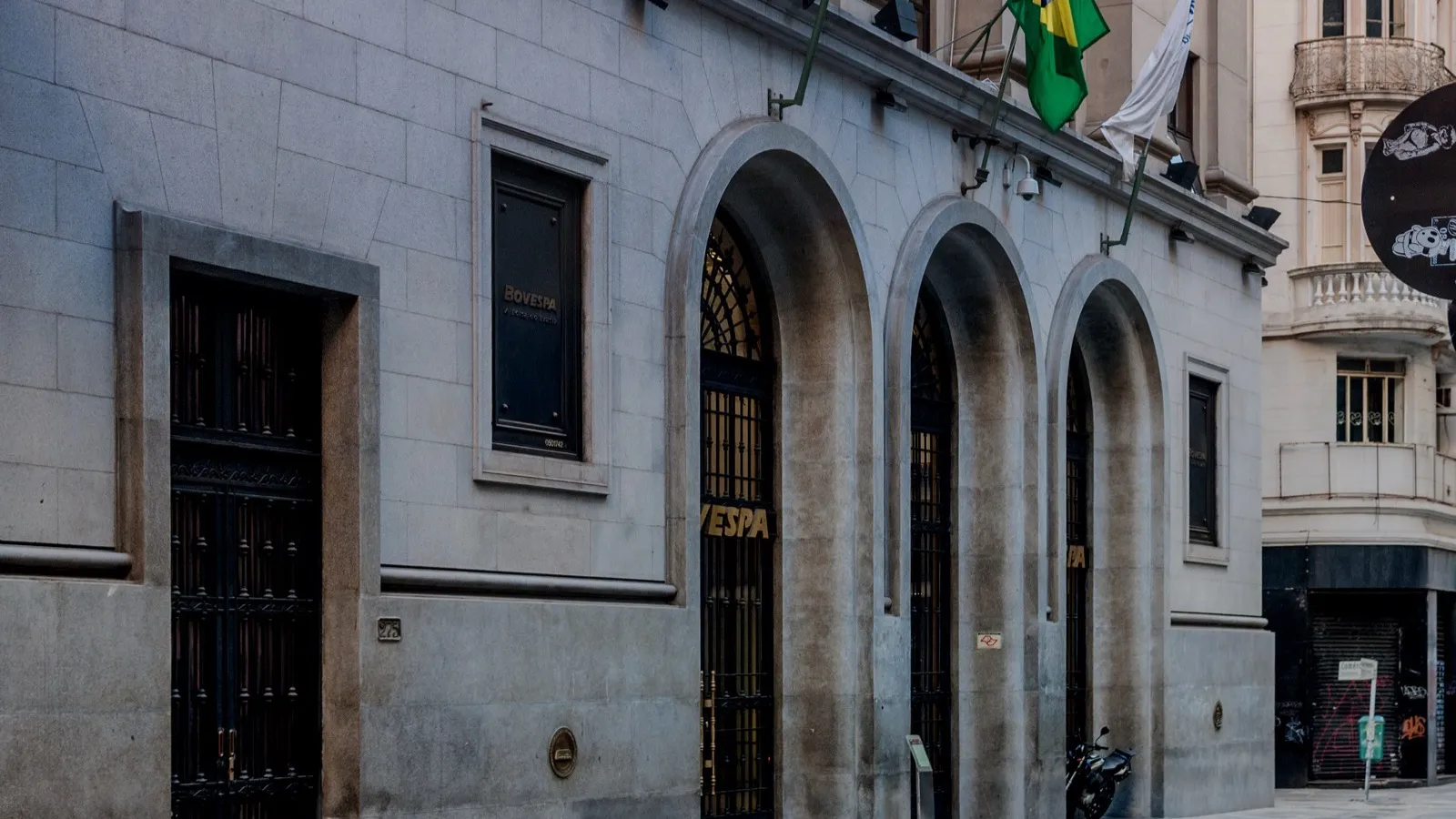 Entrada em arcos do edifício da Bovespa, com a palavra BOVESPA em letras douradas na porta e bandeira do Brasil acima