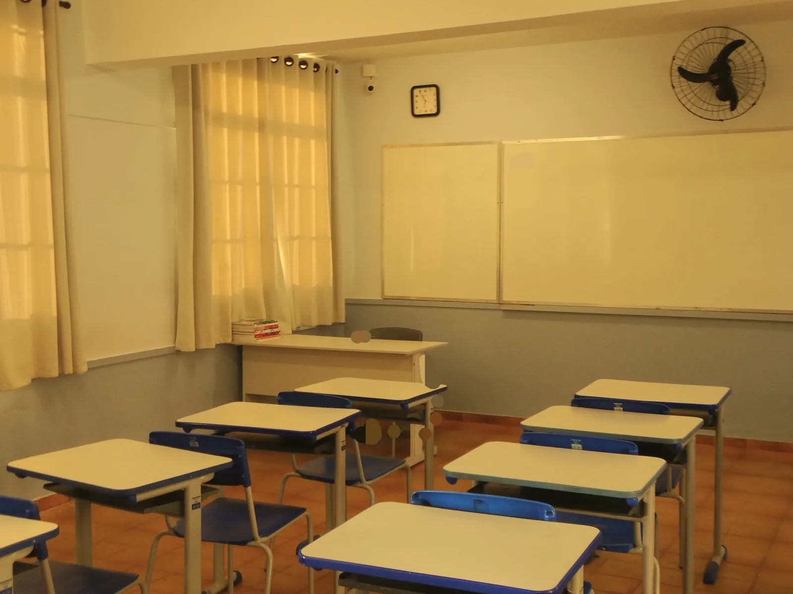 Sala de aula vazia, com carteiras azuis e brancas enfileiradas, lousa branca na parede e janela com cortinas claras