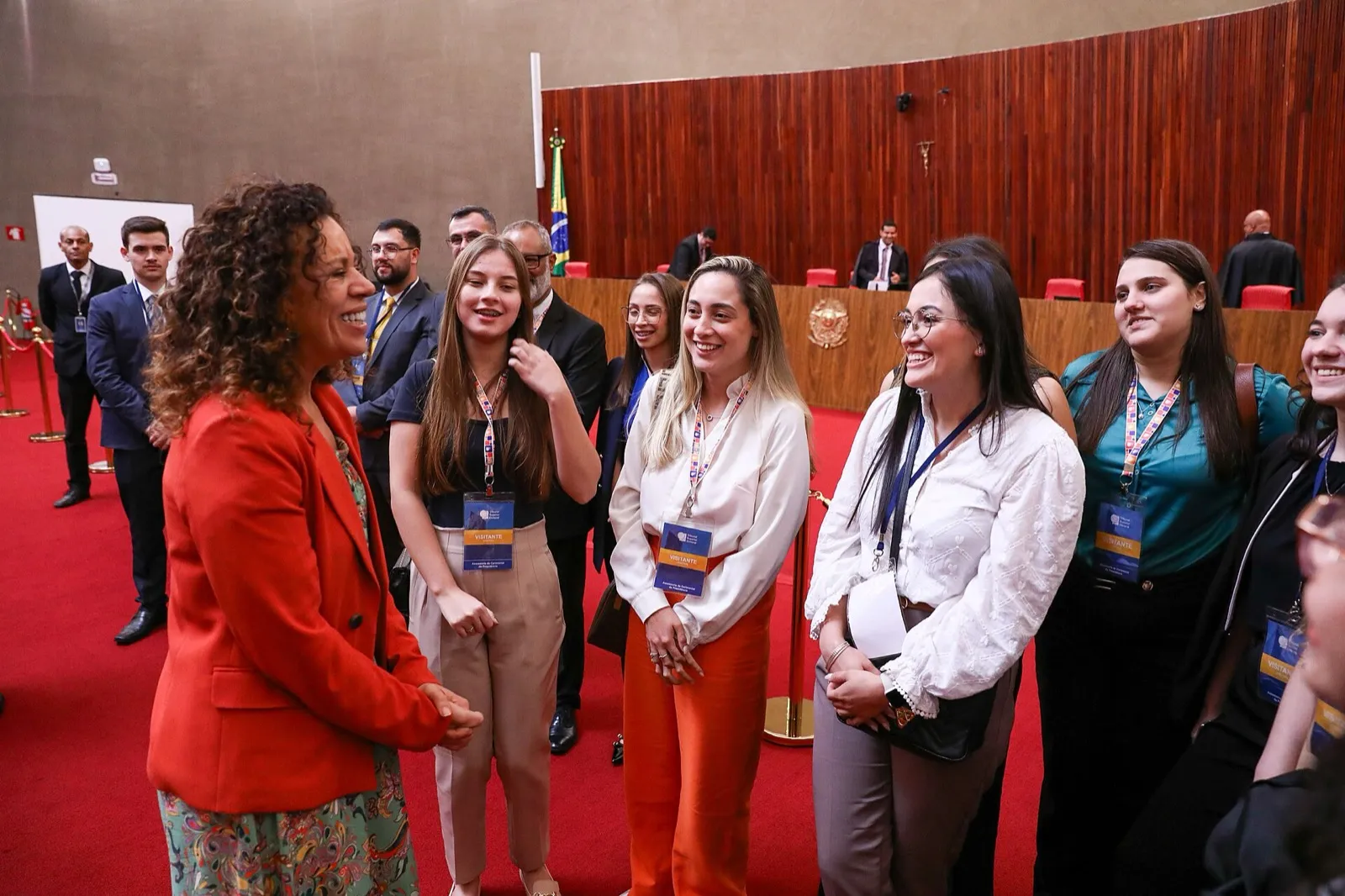 Visitantes com crachás sorriem para uma mulher de blazer vermelho no plenário do Tribunal Superior Eleitoral, com a bancada de madeira e a bandeira do Brasil ao fundo