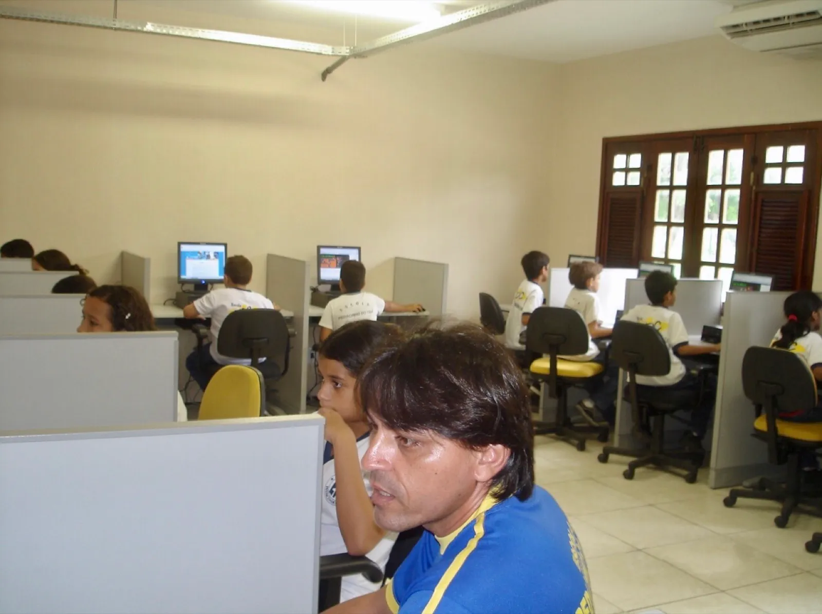 Estudantes e um professor sentados diante de computadores em laboratório escolar