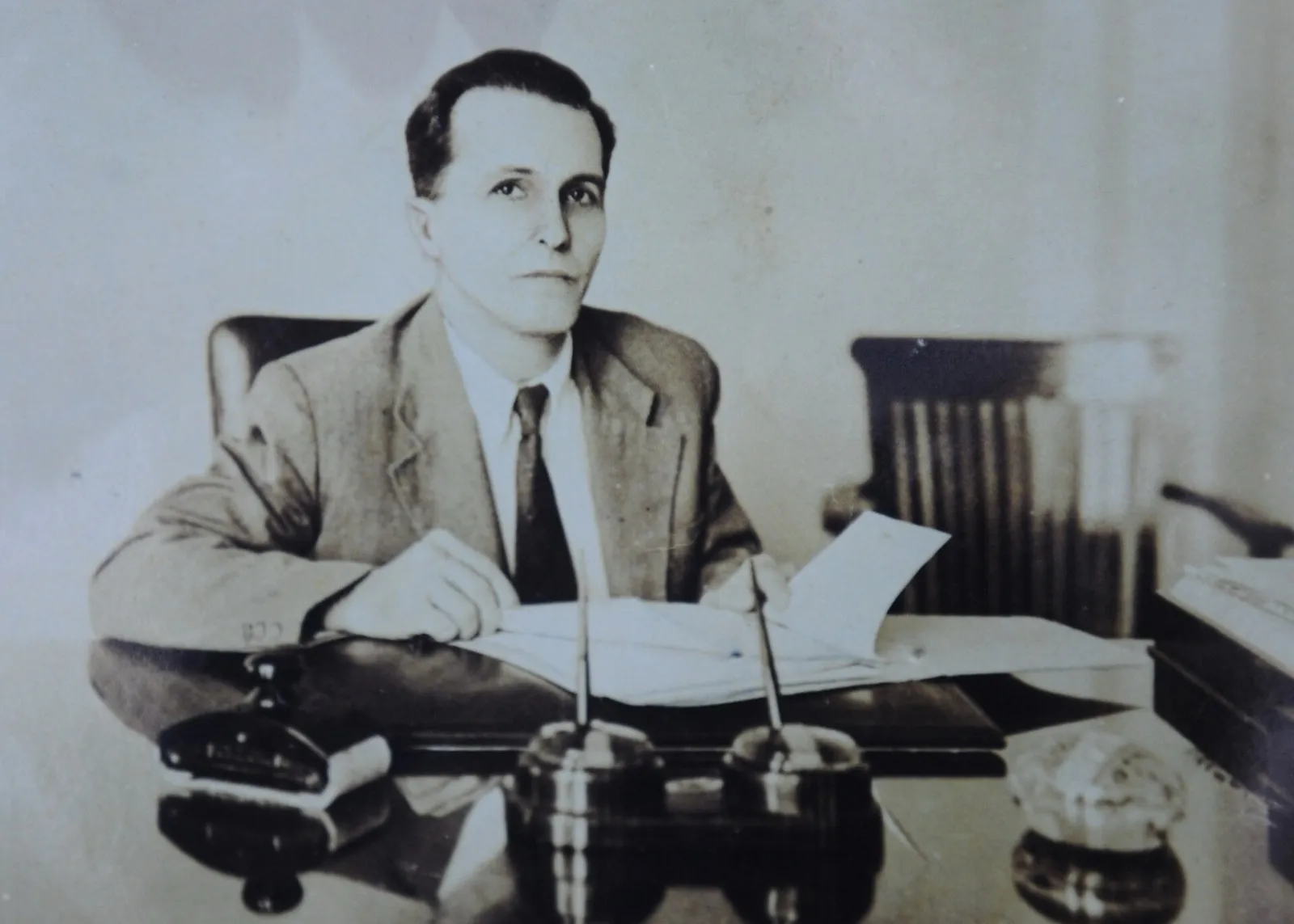 Homem de terno, sentado a uma mesa de escritório com papéis e objetos de mesa, em fotografia em tons sépia