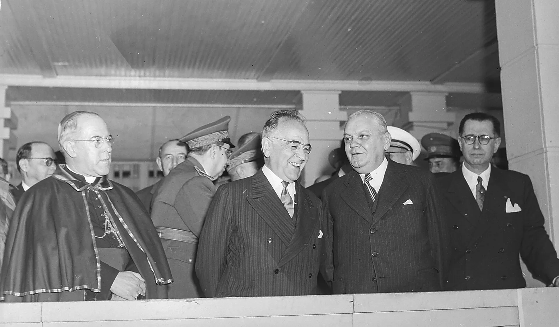 Getúlio Vargas sorri em uma tribuna ao lado de um bispo, de autoridades civis e de militares, em fotografia em preto e branco