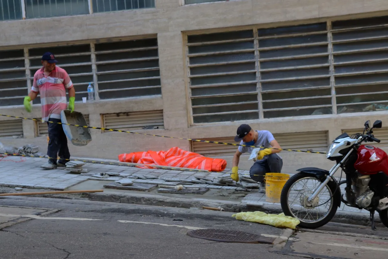 Dois trabalhadores de luvas reformam uma calçada ao lado de uma fachada, com uma moto vermelha estacionada à direita