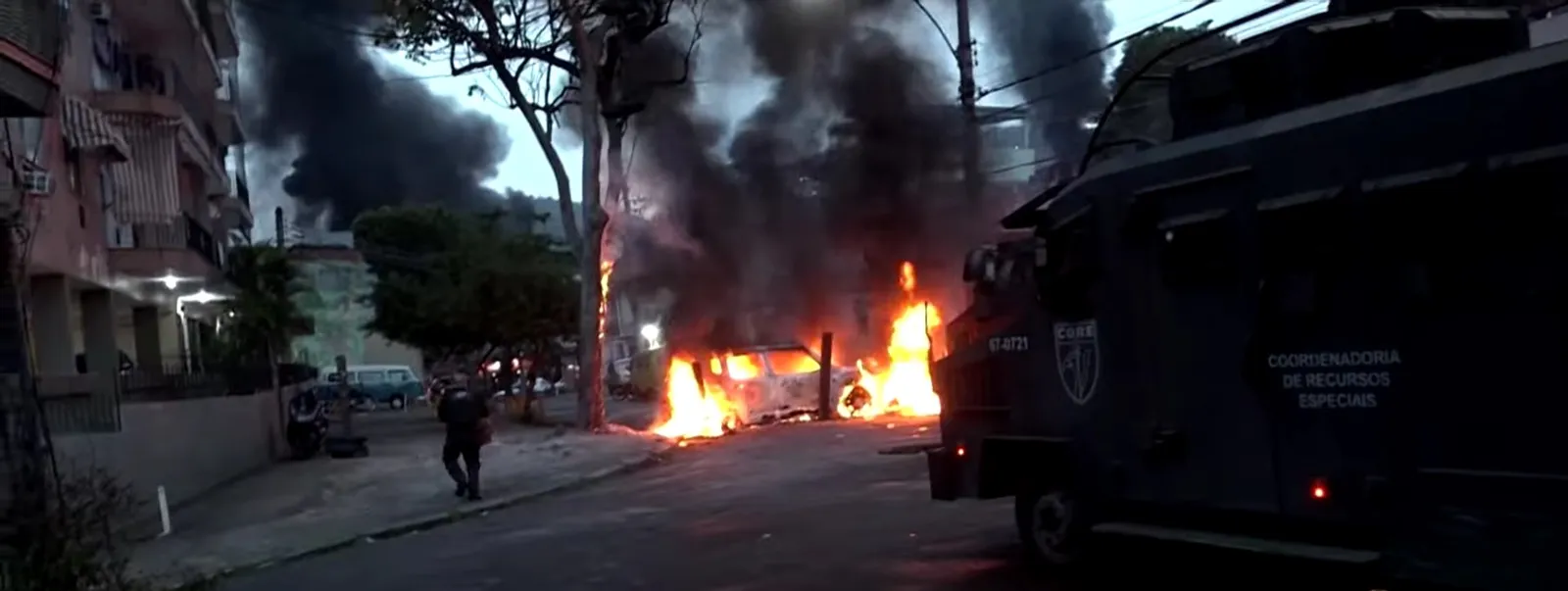 Veículo blindado da polícia diante de um veículo em chamas durante a Operação Contenção