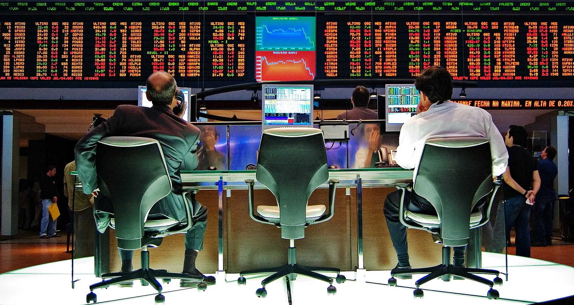 Dois operadores de costas, sentados diante de um painel com cotações de ações, em um pregão da Bovespa