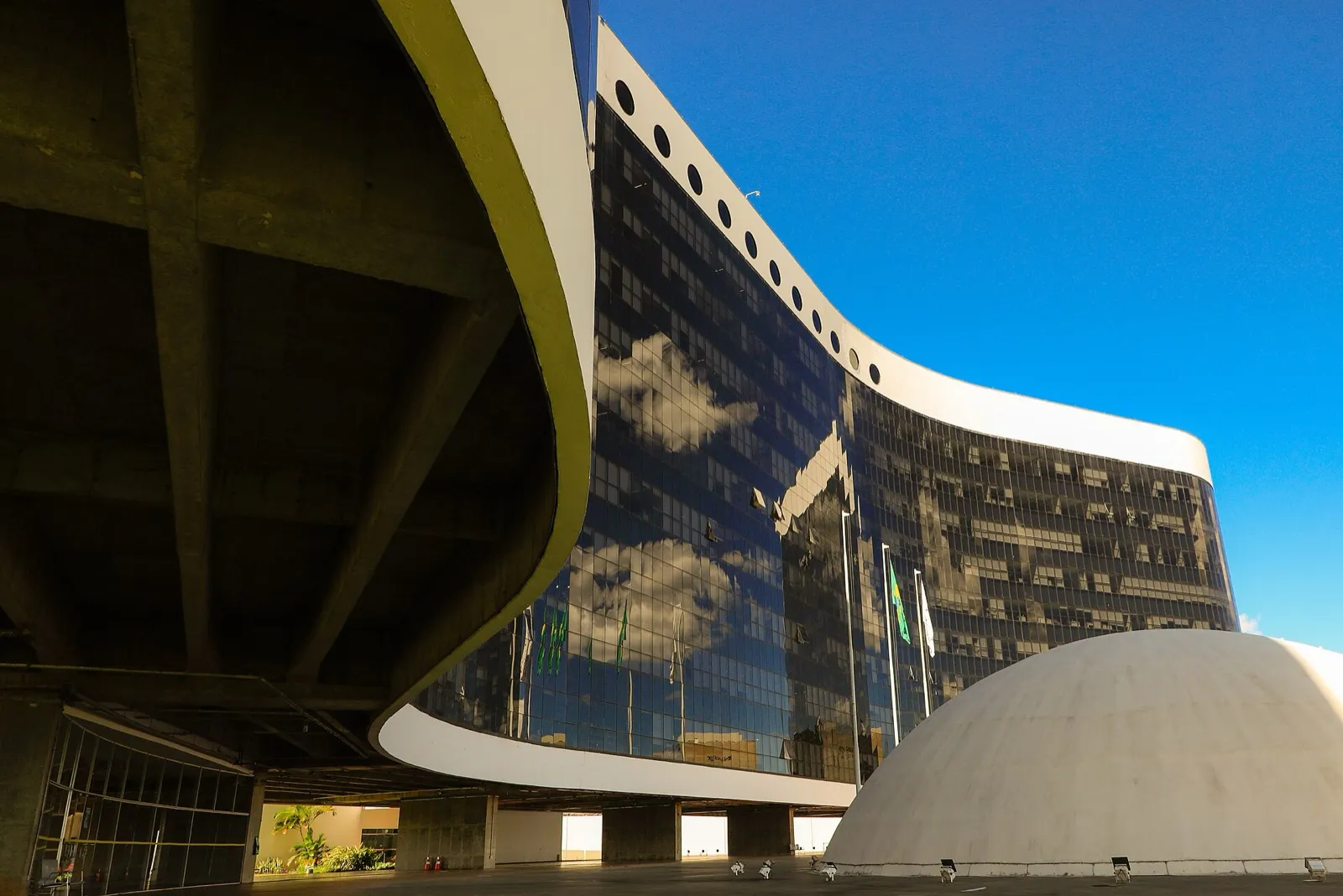 Fachada curva de vidro escuro e concreto da sede do Tribunal Superior Eleitoral, em Brasília, com cúpula branca em primeiro plano e bandeiras do Brasil ao lado