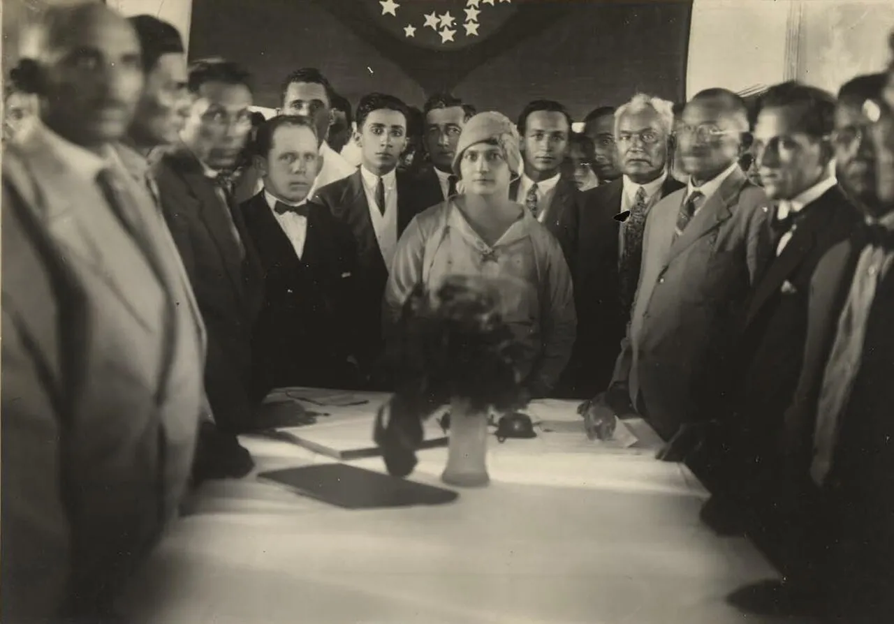 Mulher de chapéu ao centro, cercada por homens de terno diante de uma mesa e de uma bandeira, em fotografia antiga
